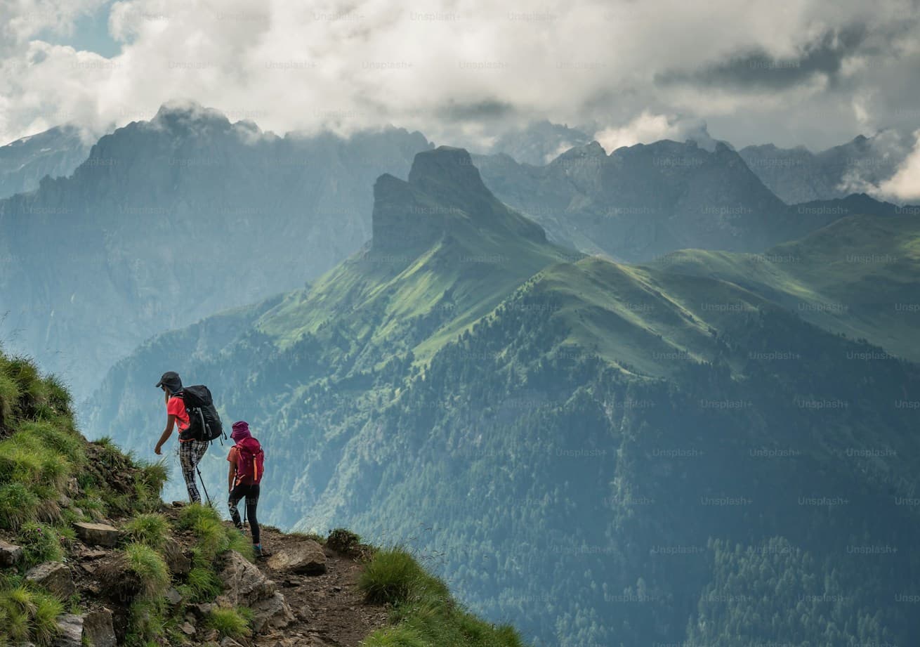 Two hikers on a ridge trail above green mountain valleys